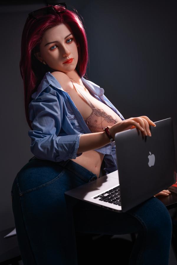 Bubbly D-cup hybrid sex doll leaning over a laptop at a desk in an open denim shirt